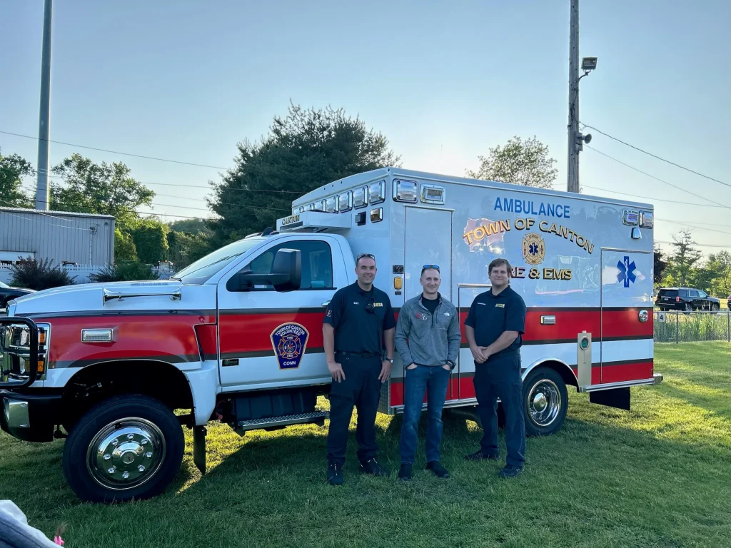 Three people stand smiling in front of a red and white ambulance labeled "Town of Canton Fire & EMS," parked on grass with trees and a sunny sky in the background.