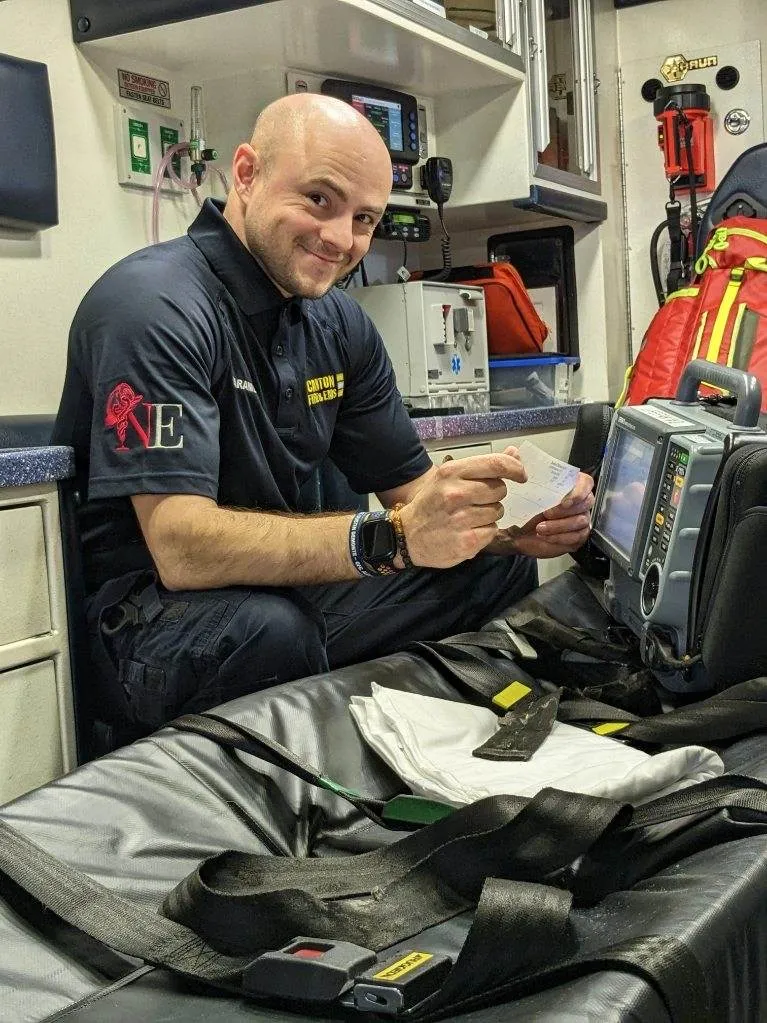 A paramedic sitting in an ambulance smiles while holding medical paperwork, with medical equipment and supplies visible around him.