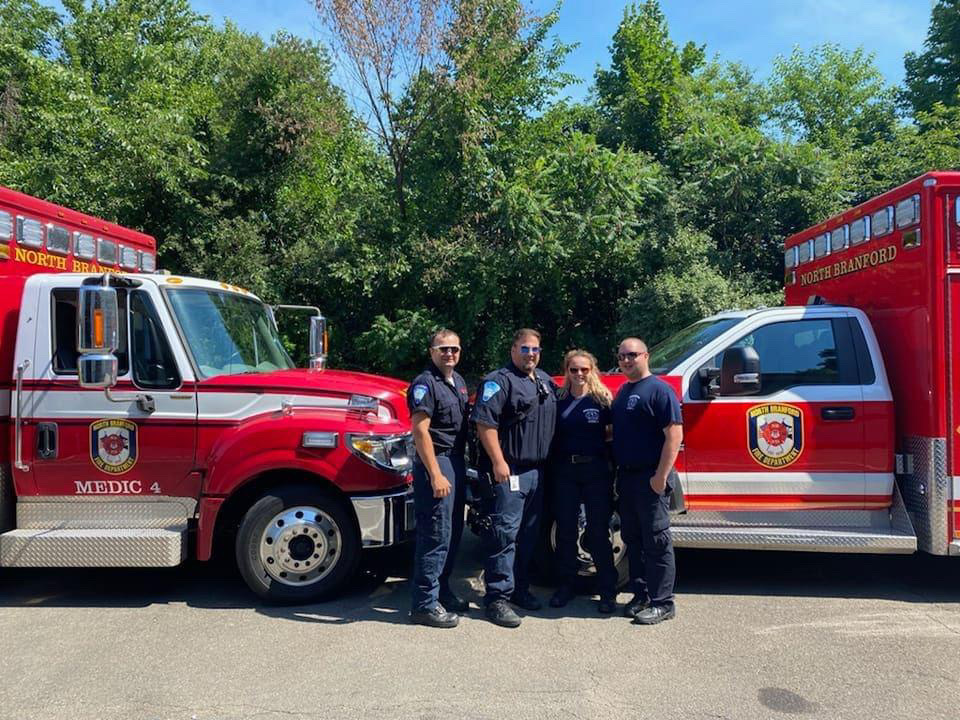 Four emergency medical personnel stand side by side in uniform between two red North Branford EMS trucks, with green trees in the background on a sunny day.