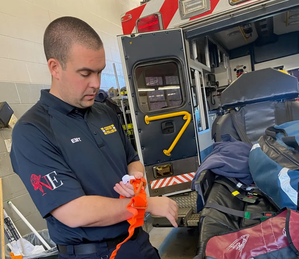 A man in an EMT uniform applies a bright orange tourniquet to his left arm inside an ambulance bay, with medical equipment and an open ambulance visible in the background.