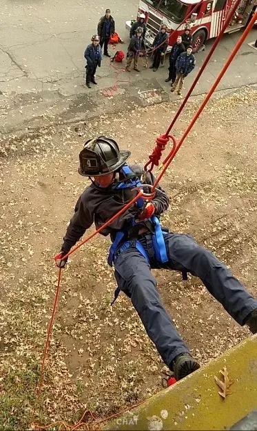 A firefighter in uniform and helmet rappels down a building using red ropes, while several people and a fire truck are visible on the ground below. Dry leaves cover the ground.
